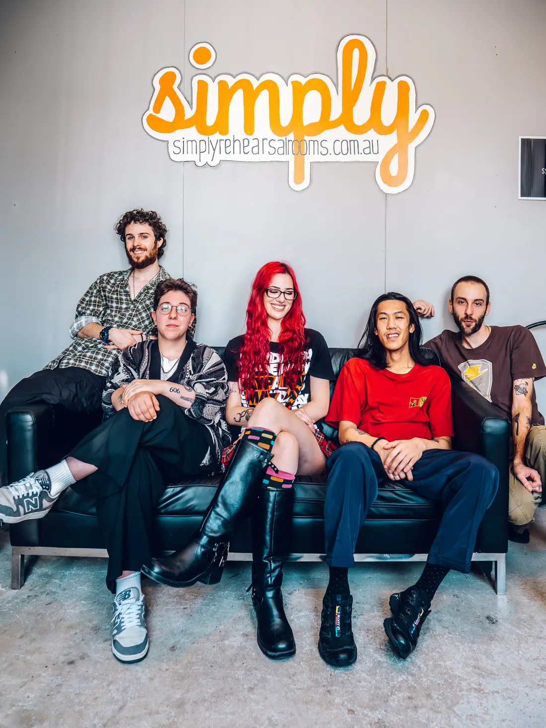 Five musicians sitting on a couch beneath the Simply Rehearsal Rooms sign
